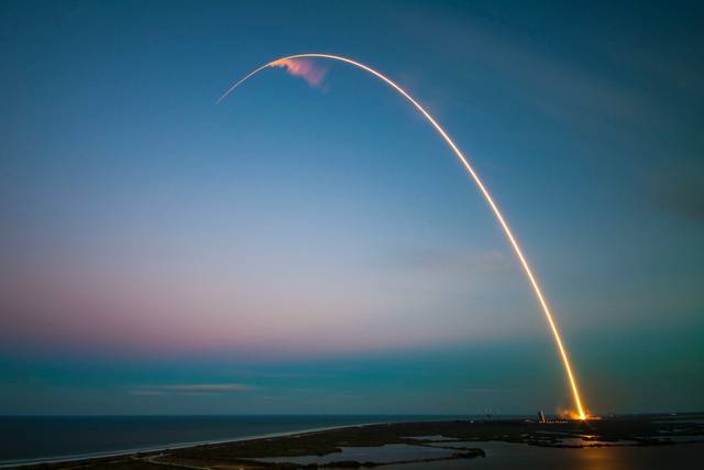 Long exposure rocket launch with arc trajectory over coastal landscape at sunset